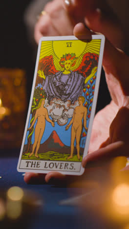 Vertical-Video-Close-Up-Of-Woman-Giving-Tarot-Card-Reading-On-Candlelit-Table-Holding-The-Lovers-Card-2